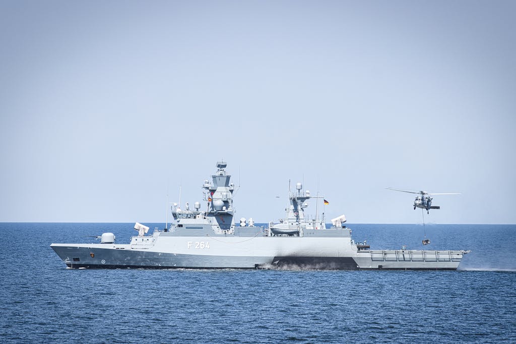 A naval frigate navigates open waters with a hovering helicopter, showcasing maritime operations.
