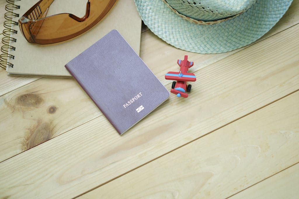 Flat lay of travel essentials including passport, notebook, toy airplane, sunglasses, and straw hat.